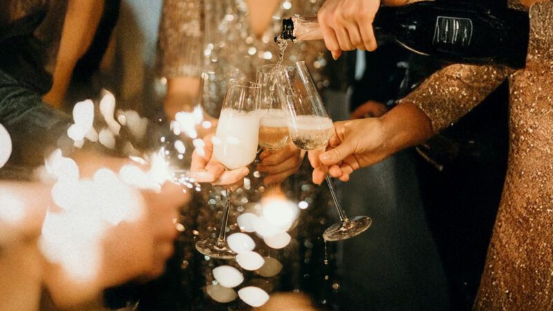 Des mains élégantes trinquent avec des coupes de champagne lors d'une soirée festive, éclairée par des lumières scintillantes et des étincelles de bonheur. Une ambiance chic et glamour qui évoque les célébrations du Nouvel An.