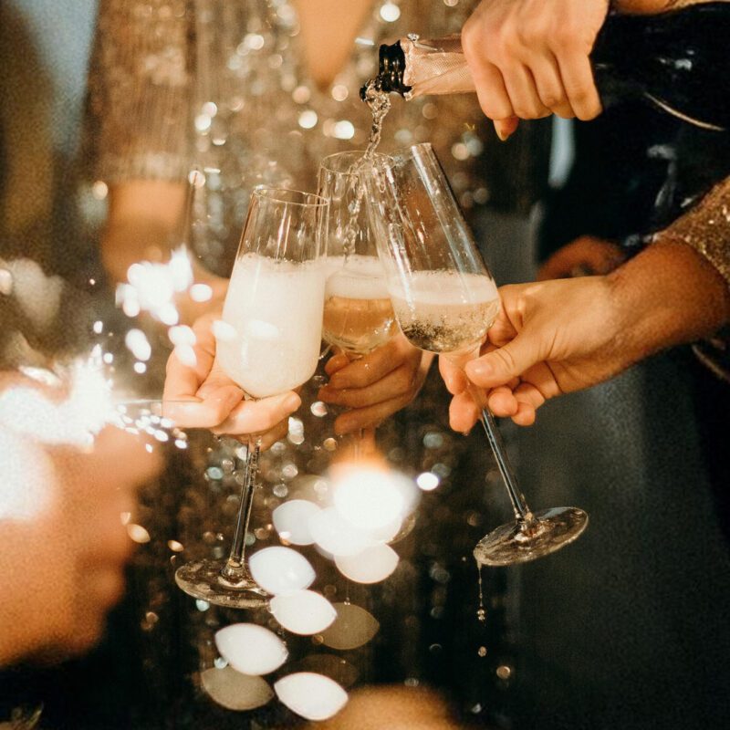 Des mains élégantes trinquent avec des coupes de champagne lors d'une soirée festive, éclairée par des lumières scintillantes et des étincelles de bonheur. Une ambiance chic et glamour qui évoque les célébrations du Nouvel An.