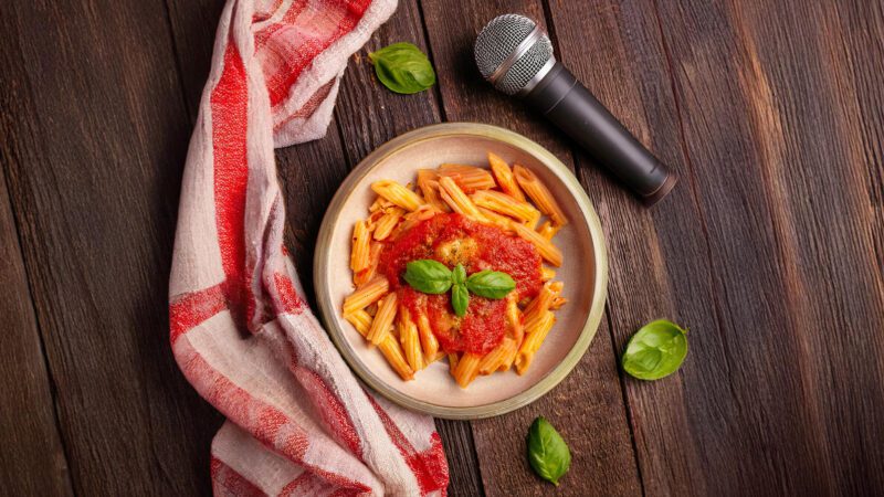 Vue plongeante sur une table en bois avec un plat de pâtes sauce tomate, un micro et un linge de cuisine.